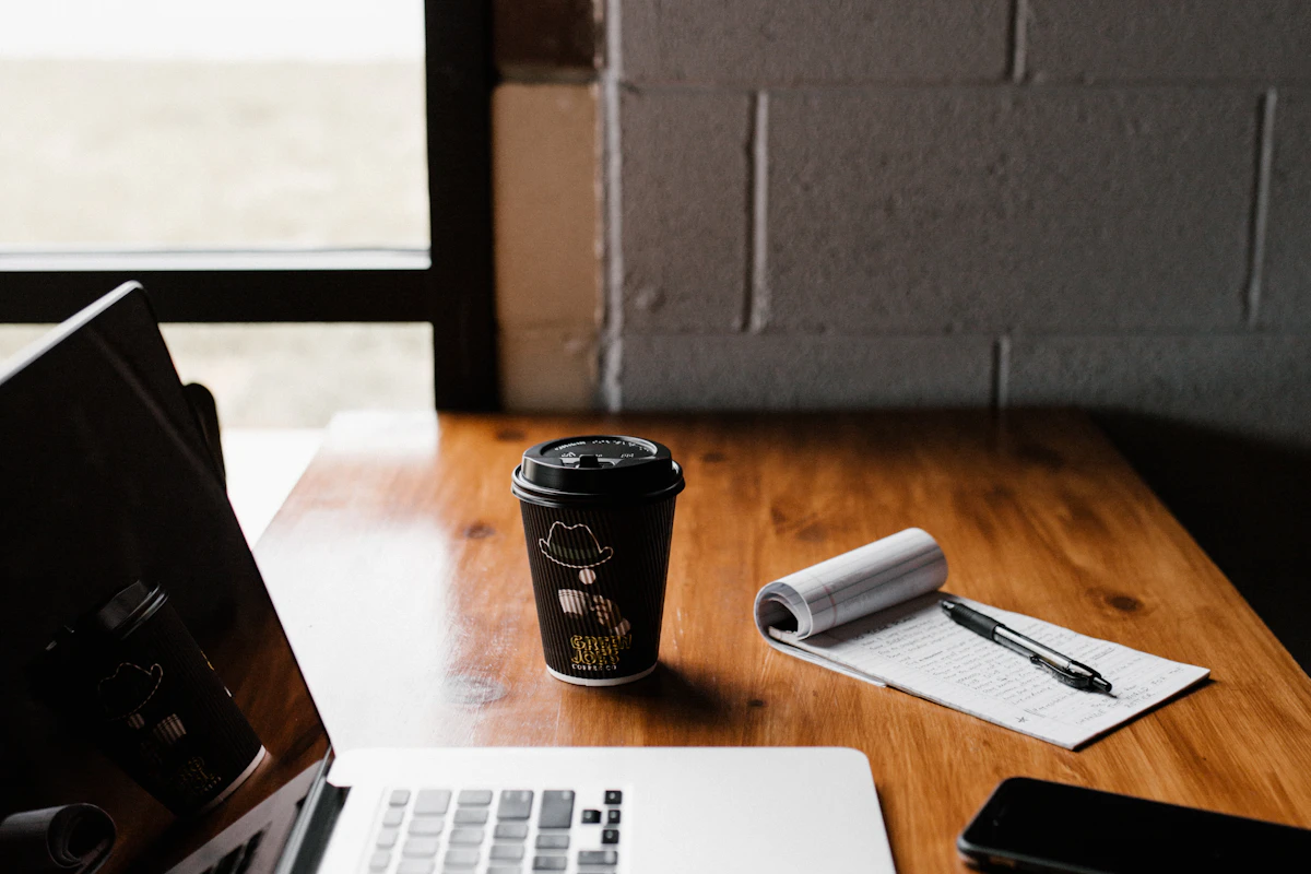 A laptop, handwritten notebook, pen, phone, and coffee cup on a wooden workspace table.